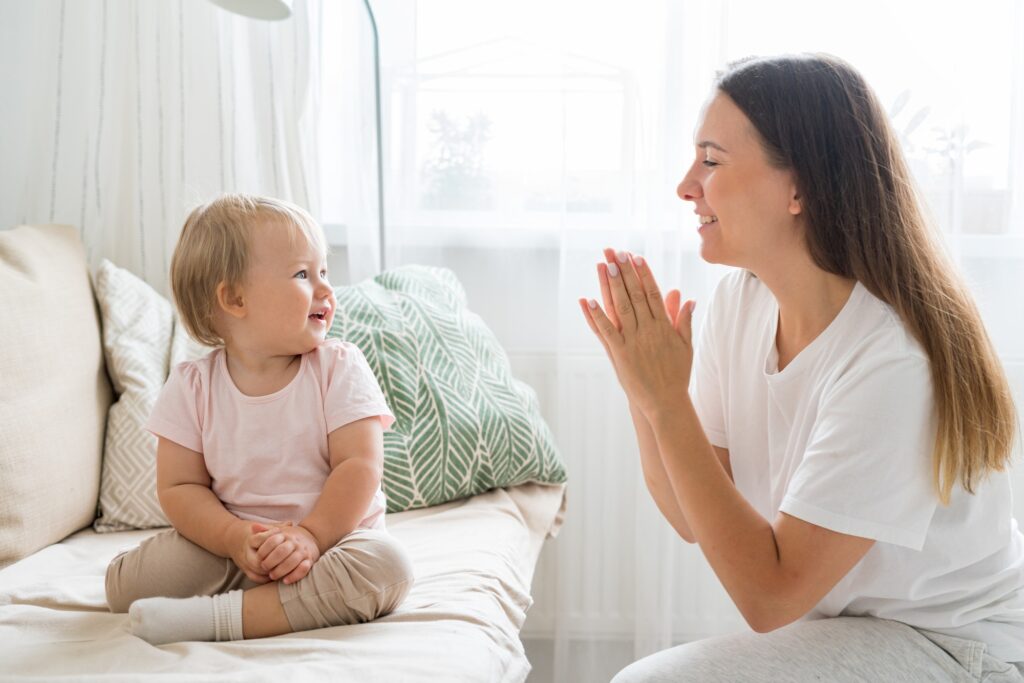 smiling mother clapping and looking at happy toddler sitting on couch in living room